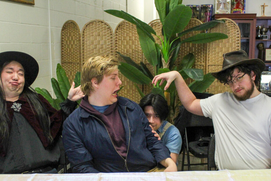 A group of four people sitting behind a table, playfully posing with exaggerated expressions. The person on the left is making a funny face, while the person on the right in a cowboy hat mimics a monster claw with his hand over another person's head.