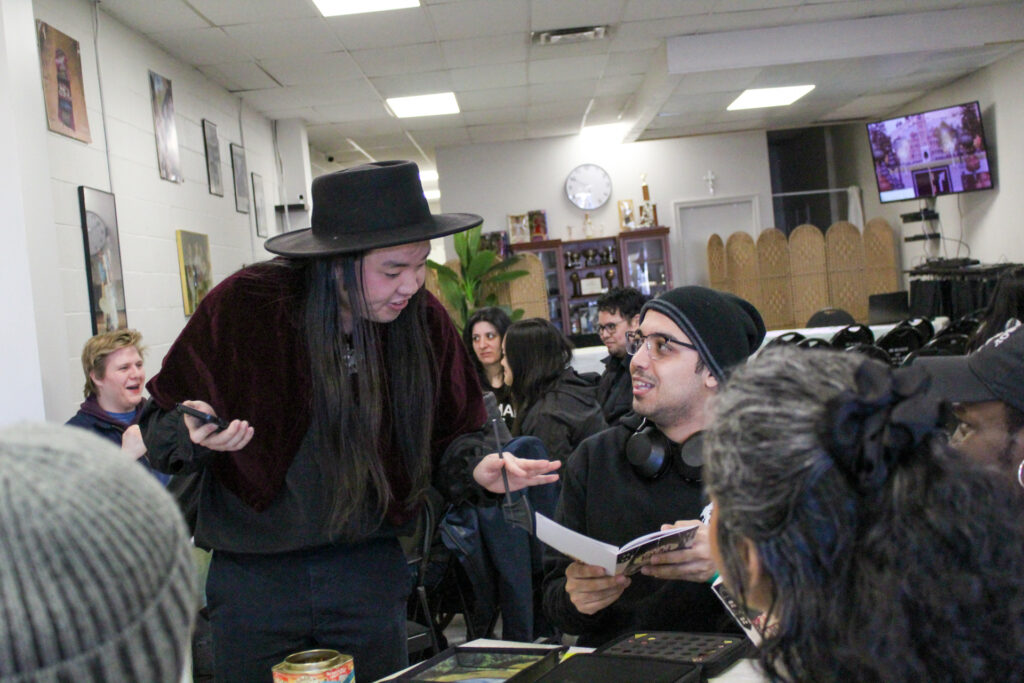 A person in a black wide-brimmed hat and maroon velvet cape stands at a table, interacting with a seated player who is holding open a small game booklet during a tabletop game session.