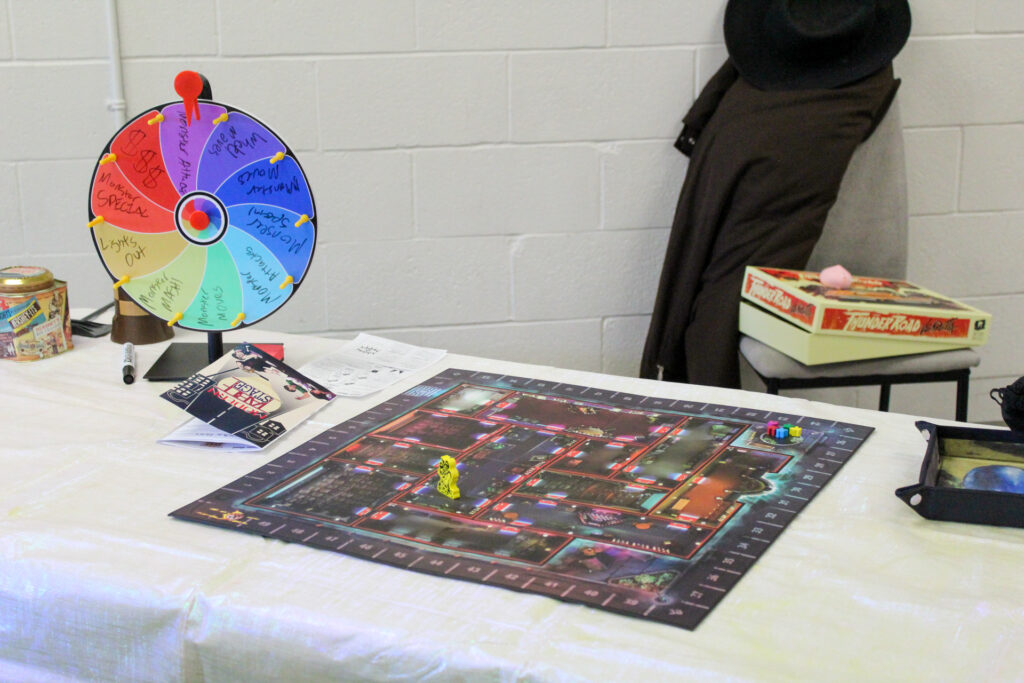 A tabletop board game set up on a white table. It features a dark, illustrated mansion floor plan map, a colourful spinning prize wheel with handwritten text options, and game pieces.