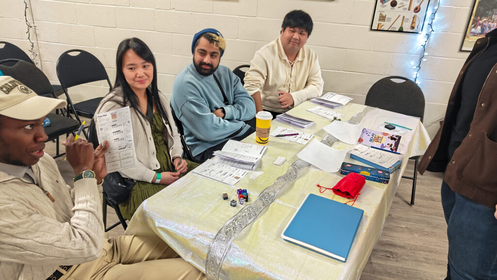 A group of five people gathered around a long table playing a tabletop game. One player in the foreground holds up a character sheet while others look on, with dice, character sheets, and notebooks scattered across the table.