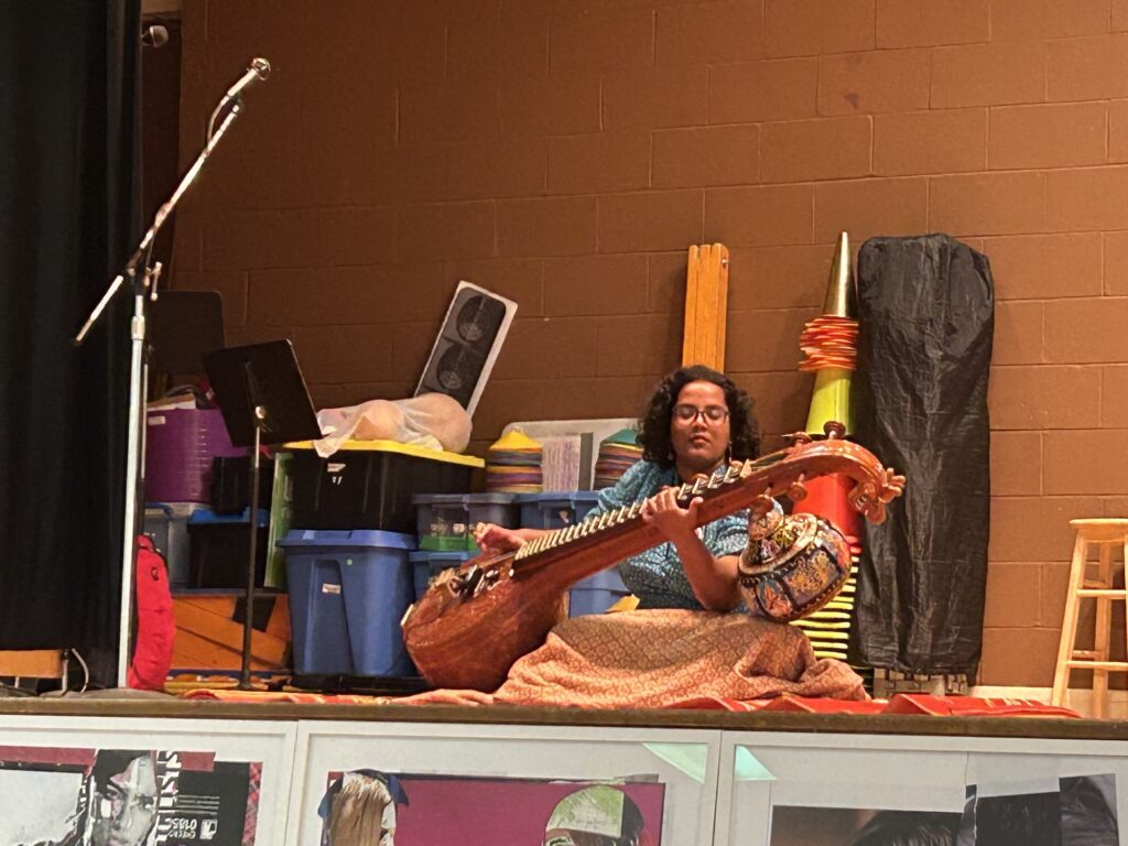 A close-up shot of a person sitting cross-legged on a stage, playing a large, ornate stringed instrument called a veena. They are wearing glasses and a blue patterned top. The background is a brown brick wall.