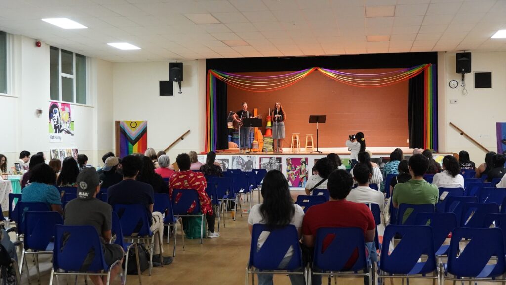 A wide view of an auditorium looking toward a stage decorated with rainbow streamers. Two musicians, one with a guitar, perform on stage under warm lighting. An audience sits in blue chairs, facing the stage from behind. Artwork is displayed along the front base of the stage.