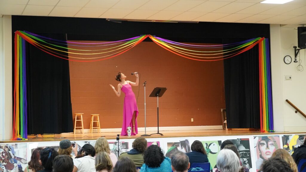 A performer in a long pink dress sings into a microphone on a stage decorated with rainbow fabric streamers draped across the top and sides. The stage has a brown brick backdrop, two wooden stools, and a music stand. An audience sits in front of the stage, viewed from behind.
