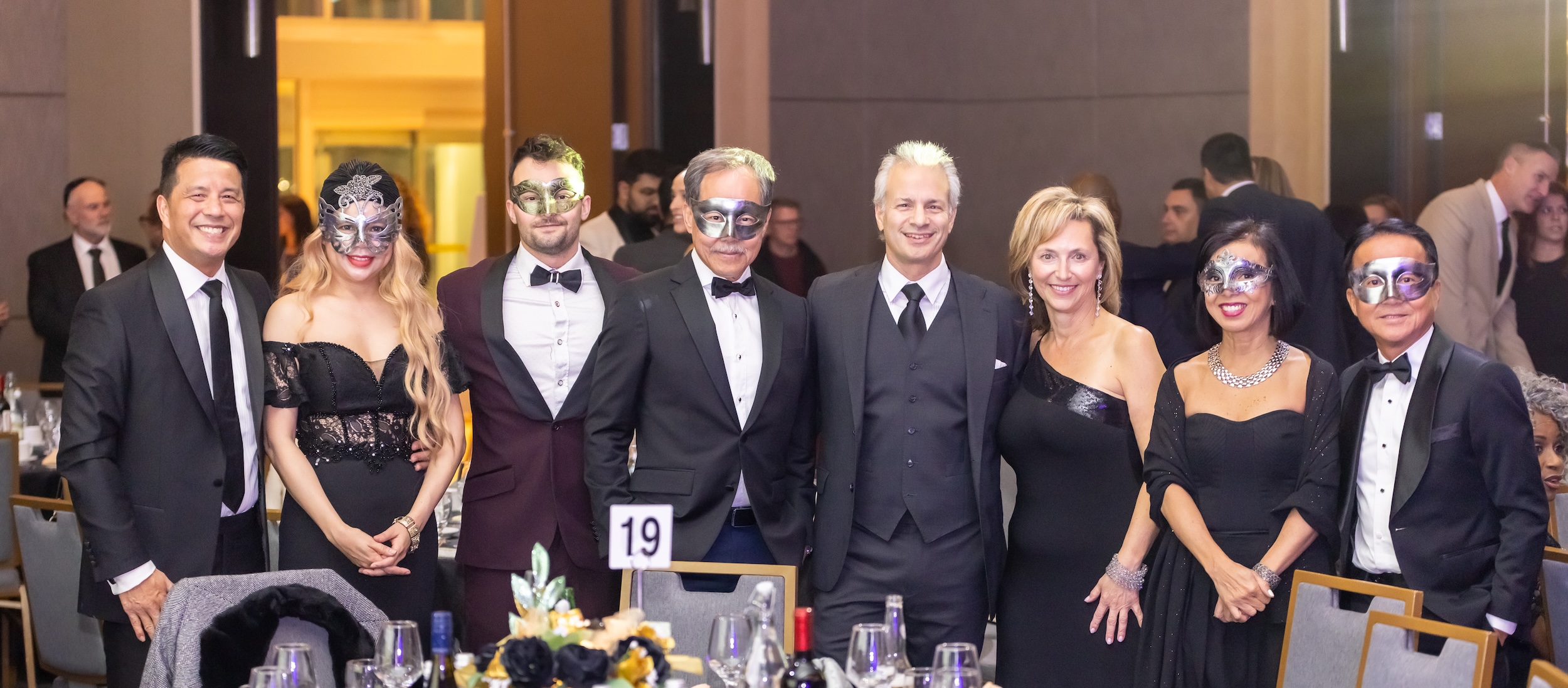 A group of eight guests in formal black-tie attire and decorative masquerade eye masks smile together for a portrait around Table 19 at a gala event.