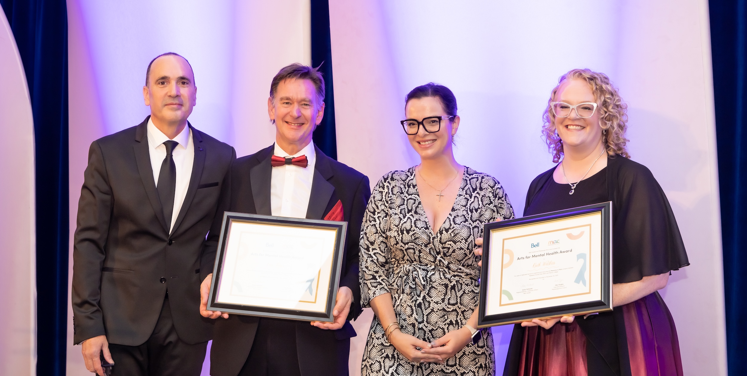Four people posing on stage at the 2025 MACsquerade Glam Ball. The two individuals in the center and on the right are holding framed "Arts for Mental Health Award" certificates.