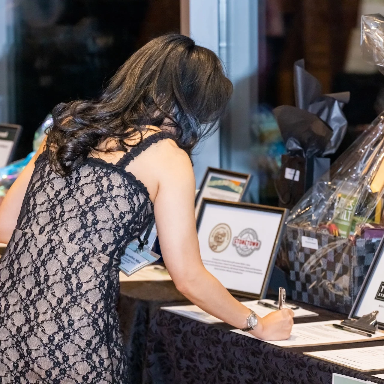 A woman is seen from behind writing a bid on an auction sheet in front of a table full of framed prizes and gift baskets.