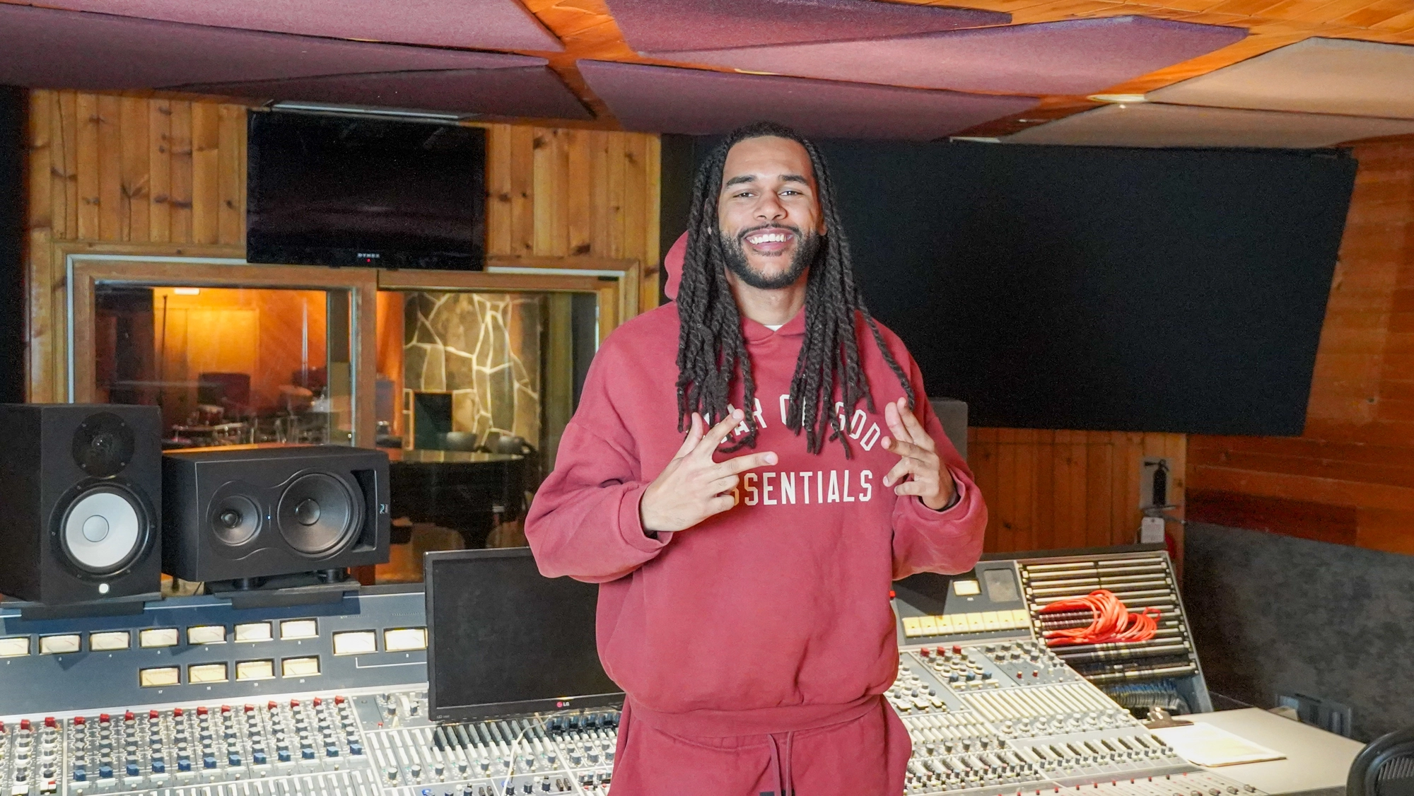 Neddy stands against a mixing table at a recording studio. He smiles in a red hoodie, and holds up two peace signs with his fingers.