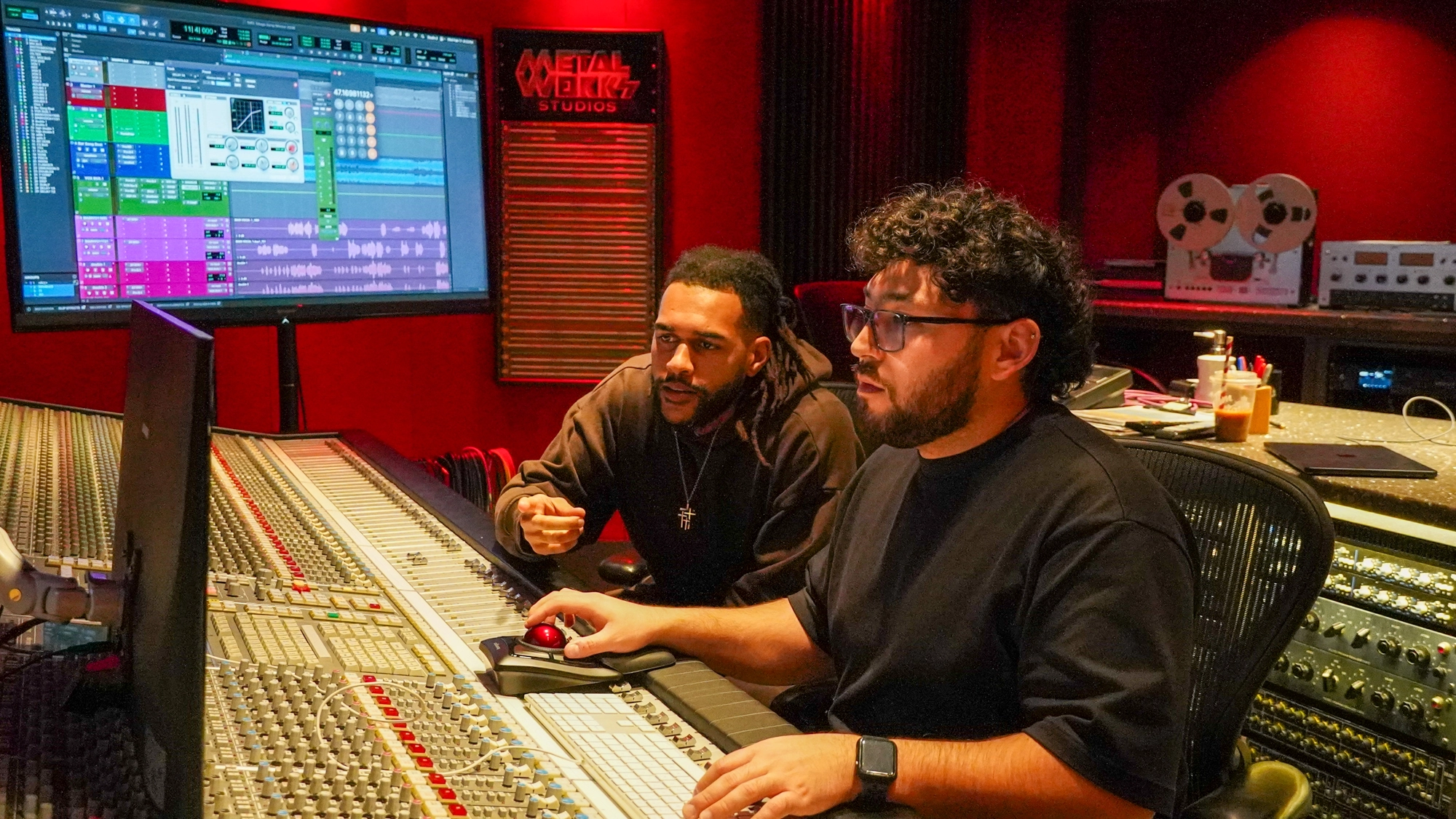 Two people collaborate at a large mixing console in a dimly lit, red studio room labeled "Metalworks Studios."